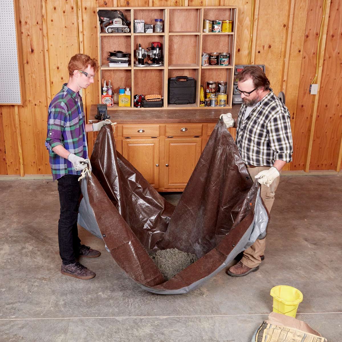 Two men lift a large tarp filled with materials in a wooden workshop, surrounded by shelves and cabinets containing tools and supplies.
