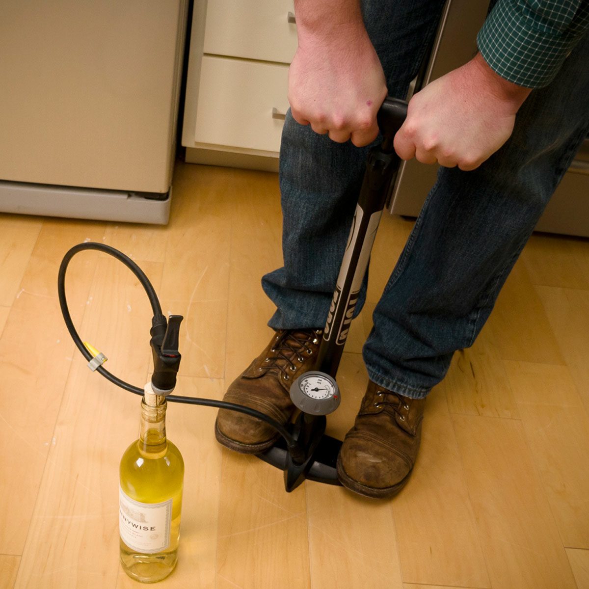 A person uses a pump with a gauge to pressurize a wine bottle in a kitchen setting, standing on a wooden floor near cabinetry.