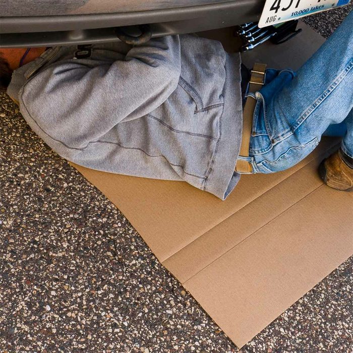 A person lies under a vehicle, inspecting it while resting on a piece of cardboard, surrounded by a gravel driveway.