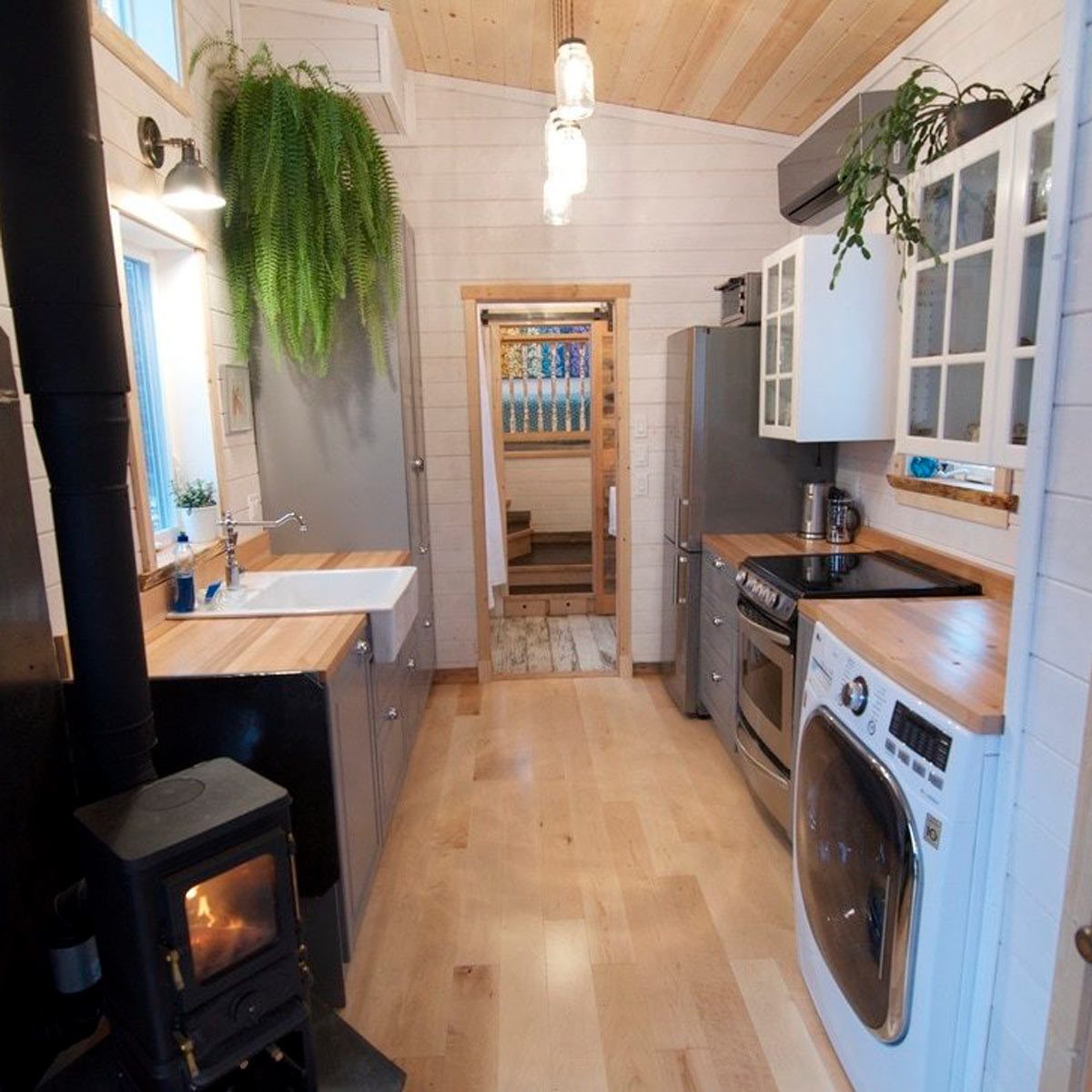 A modern kitchen contains a sink, appliances, and wooden countertops. A small wood stove emits warmth. A doorway leads to a light-filled hallway.