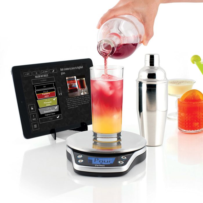 A hand pours cranberry juice into a glass on a scale, while an iPad displays cocktail instructions; a shaker and drinks sit nearby.