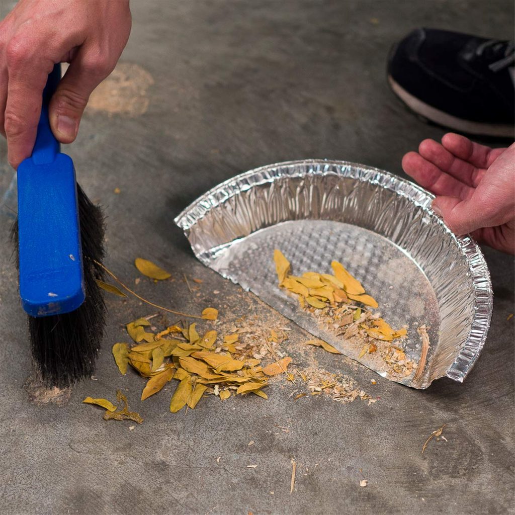 A hand holds a blue brush, sweeping debris into a metal pie dish on a gray floor, while another hand tilts the dish to catch the dirt.