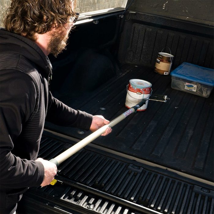 A person uses a long tool to reach a paint can in a truck bed, surrounded by other containers and tools in a well-lit environment.