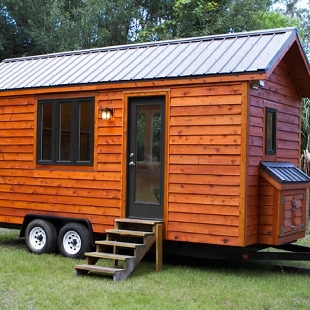 A small wooden house rests on wheels, featuring a metal roof, large windows, and a front porch with stairs, surrounded by grass and trees.