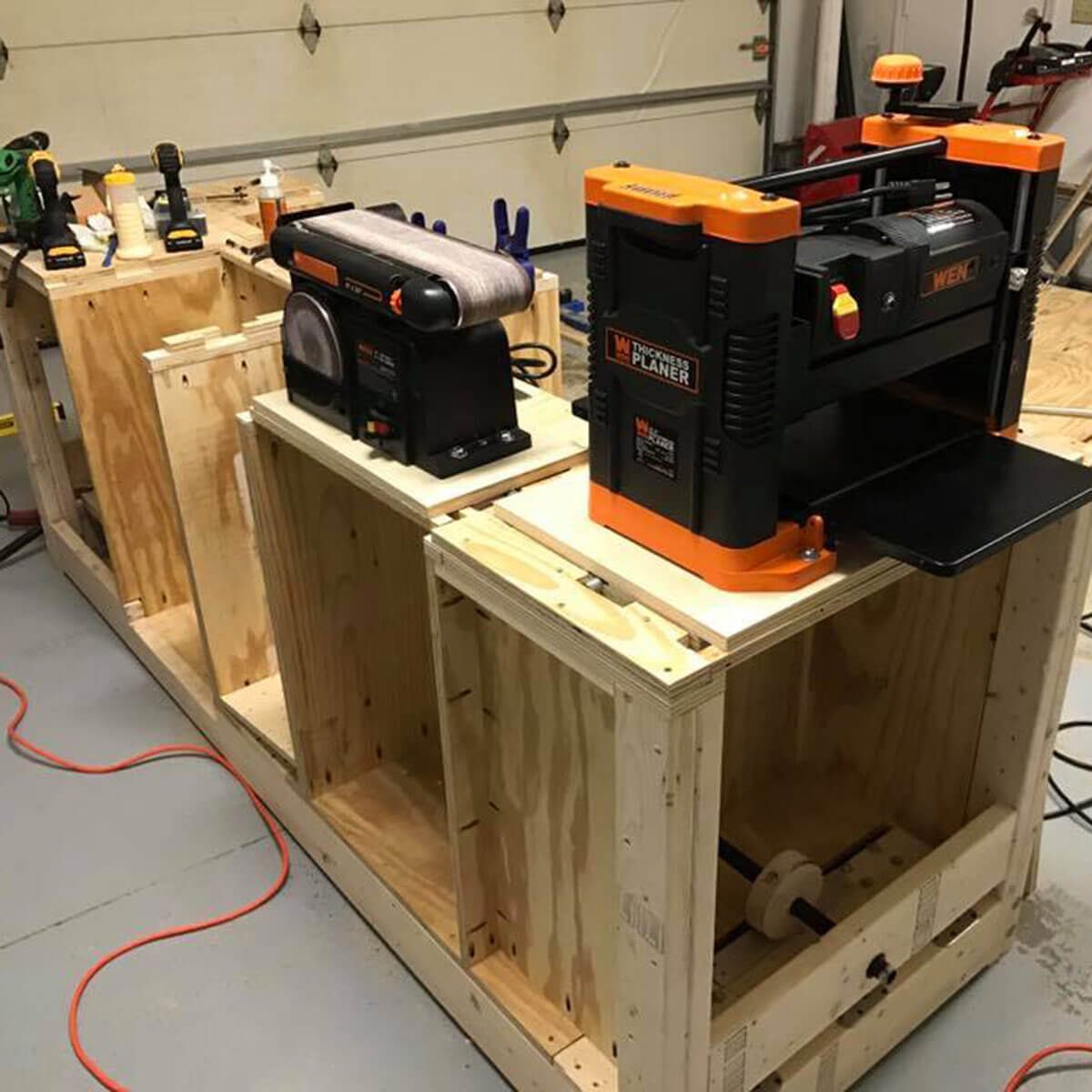 A wooden workbench showcases a thickness planer and sander, surrounded by various tools, with orange electrical cords scattered on the floor in a garage setting.