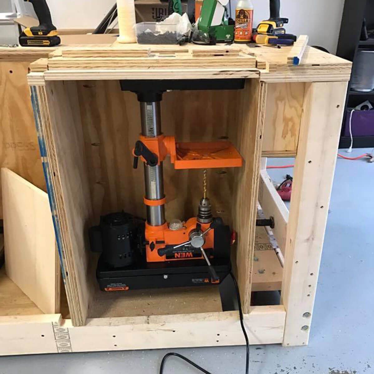 A drill press is positioned inside a wooden enclosure, actively ready for use. Surrounding tools and materials suggest an organized workspace.