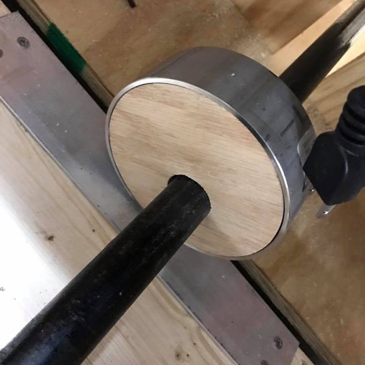 A circular wooden and metal object sits near a black rod on a workbench, suggesting it may be part of a tool or assembly.