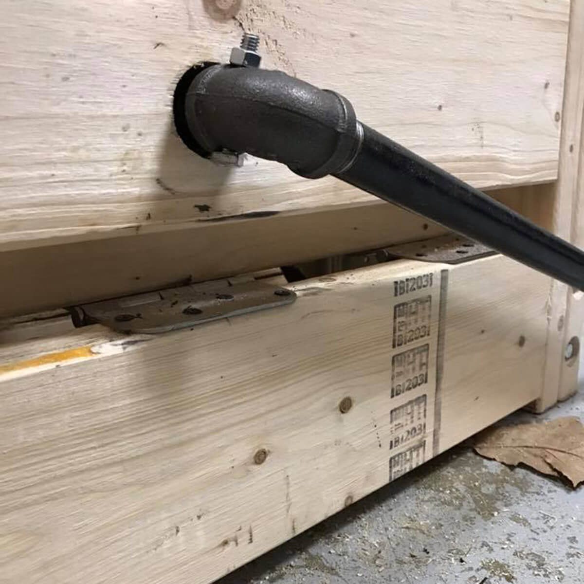 A black pipe extends through a hole in wooden planks, resting above a wooden box with visible markings, in a dimly lit, slightly untidy environment.
