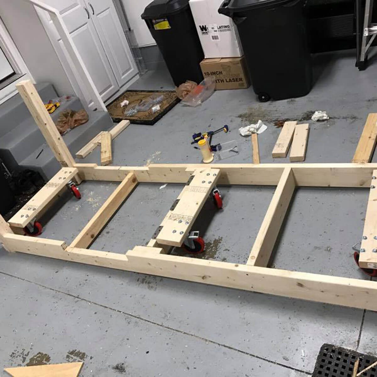 A wooden frame with wheels is assembled on a garage floor, surrounded by tools and materials, indicating a woodworking project in progress.