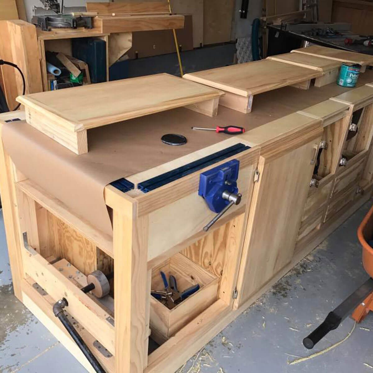 A wooden workbench with multiple drawers and shelves is being organized in a workshop, featuring tools, a vise, and a roll of paper.
