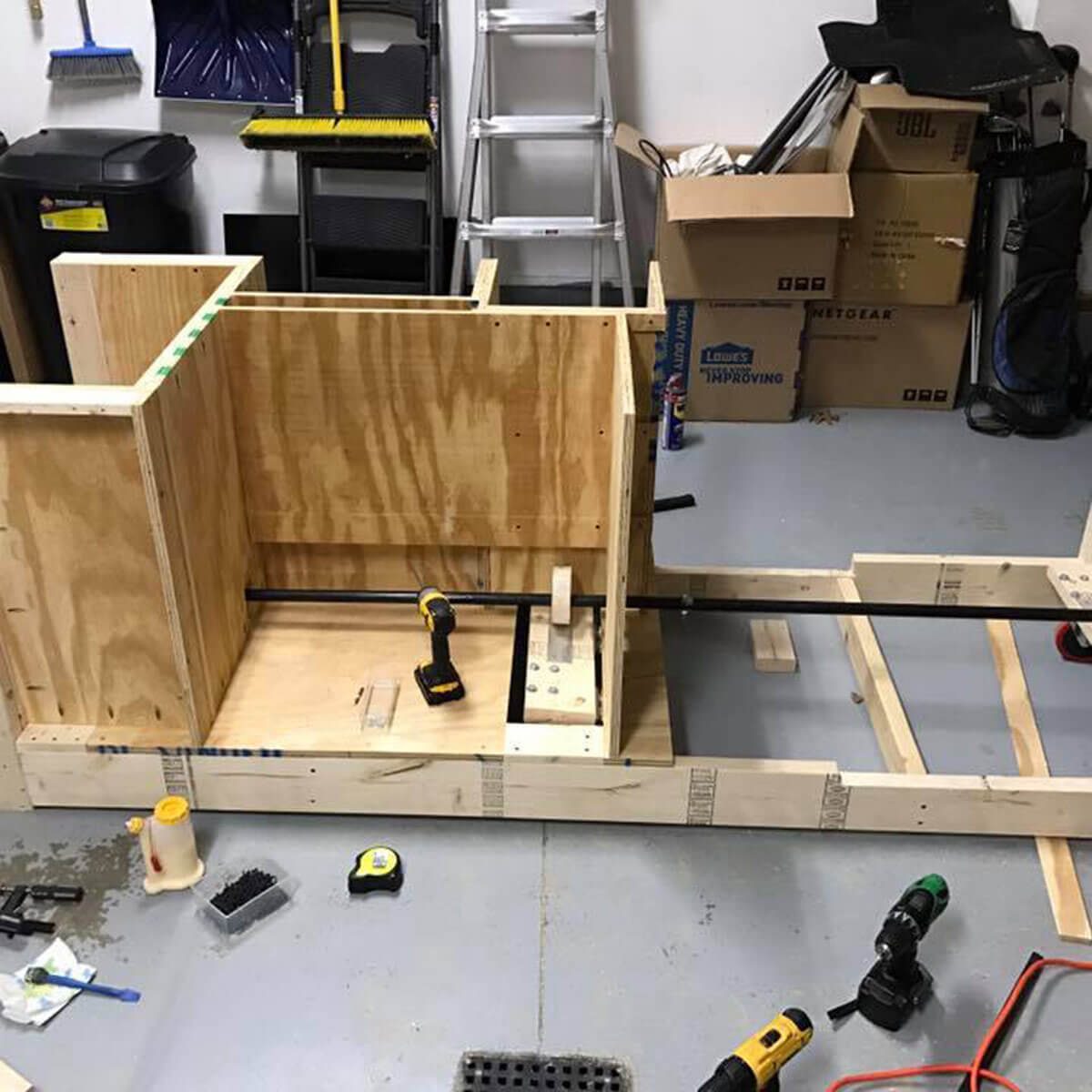 A wooden structure is being assembled on a garage floor, with tools and materials nearby, amid boxes and cleaning equipment in the background.