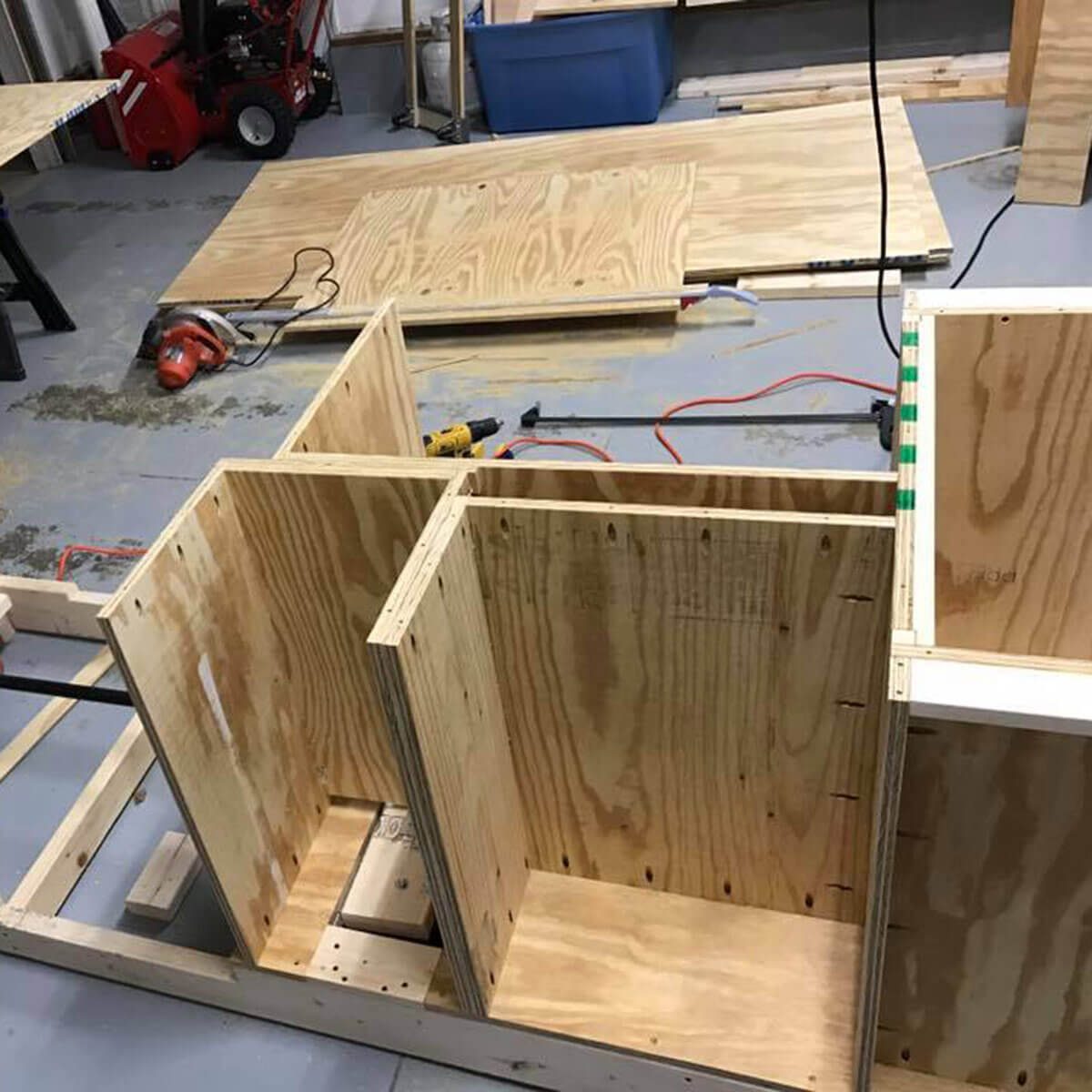 Wooden pieces are being assembled into a structure on a workshop floor, surrounded by tools and additional wooden panels. Electric cords are visible.