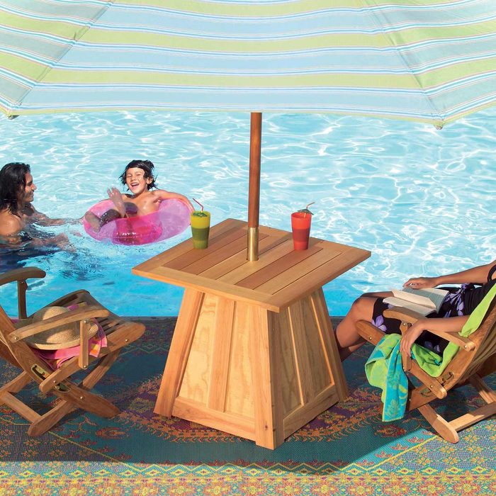 A wooden table with drinks stands under a striped umbrella beside a pool, while a child swims and two adults relax in chairs.