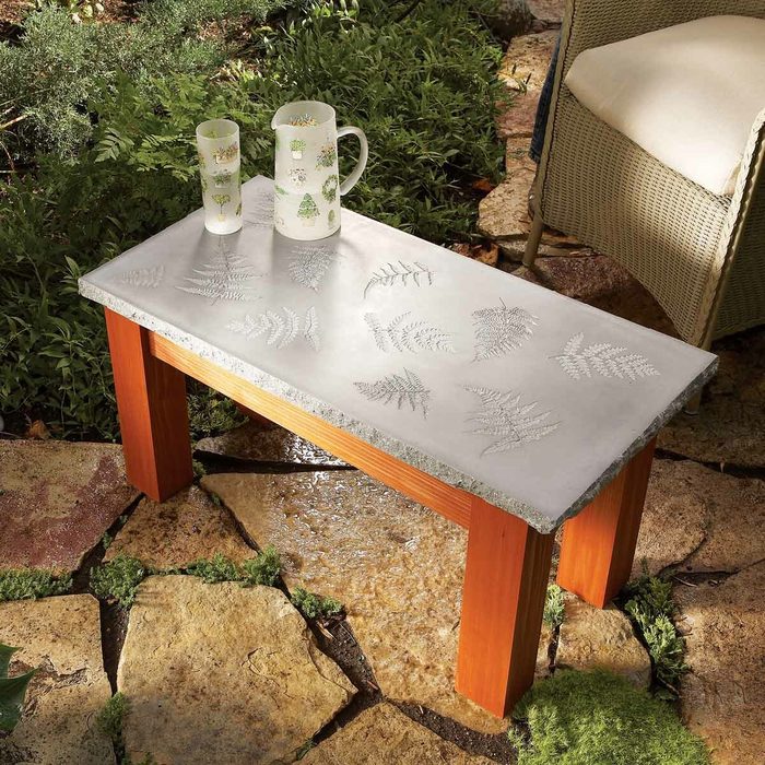 A small outdoor table with a concrete top featuring leaf patterns, supported by wooden legs. On the table are a decorative pitcher and glass. The surrounding area has a stone-tiled path and greenery.