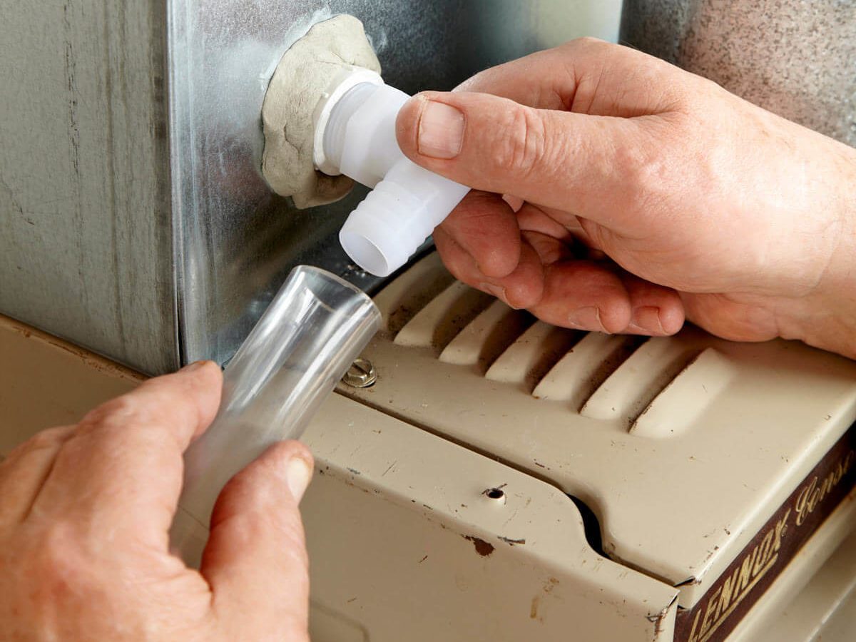 A person is attaching a clear tube to a white fitting on a furnace in a mechanical setting, using their hands to secure the connection.