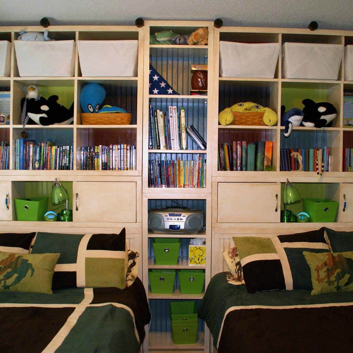 A tall shelving unit filled with books, toys, and decorative baskets stands between two neatly made beds in a brightly colored room.