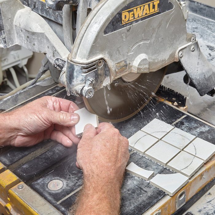 A person uses a tile saw to cut pieces of tile, with water spraying to minimize dust. The work surface is covered in tile crumbs.