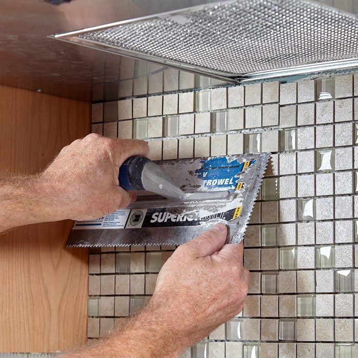 A person uses a trowel to apply adhesive to a tiled surface in a kitchen, surrounded by wood cabinetry and a metallic vent.