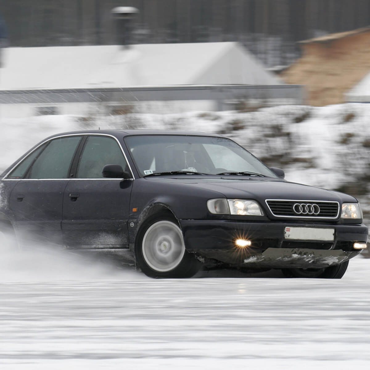 A black Audi car drifts on a snowy surface, creating a spray of snow while surrounded by a blurred winter landscape and white structures in the background.