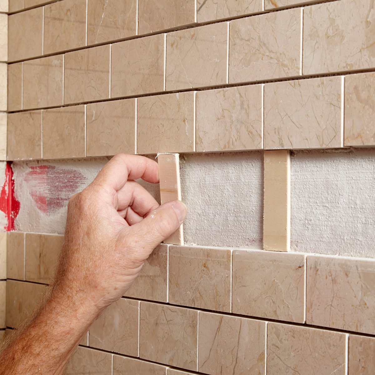 A hand positions a tile piece into a wall space among rectangular, beige tiles, showcasing a partially finished tiling project in a construction setting.