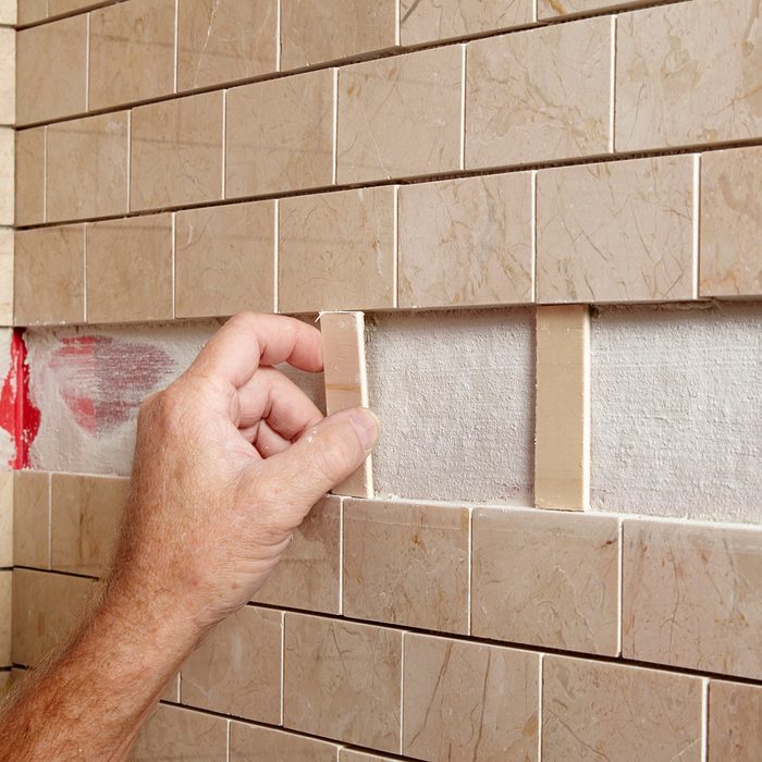 A hand positions a tile piece into a wall space among rectangular, beige tiles, showcasing a partially finished tiling project in a construction setting.
