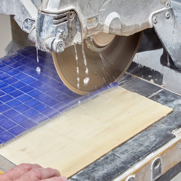 A circular saw cuts through a wooden board, spraying water onto the surface, while a blue tiled surface serves as a backdrop in a workshop setting.