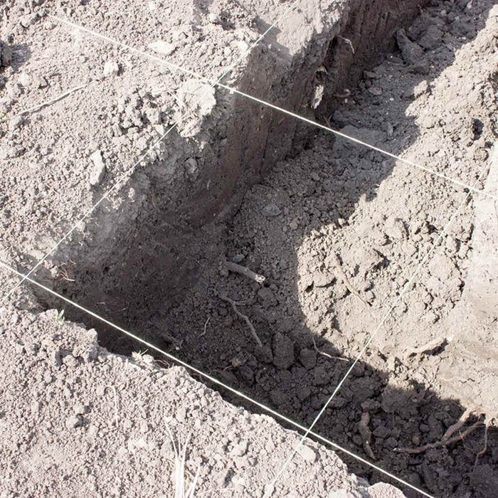 A rectangular trench is dug into loose, dry soil, with strings marking its dimensions, surrounded by excavated earth and sparse vegetation.