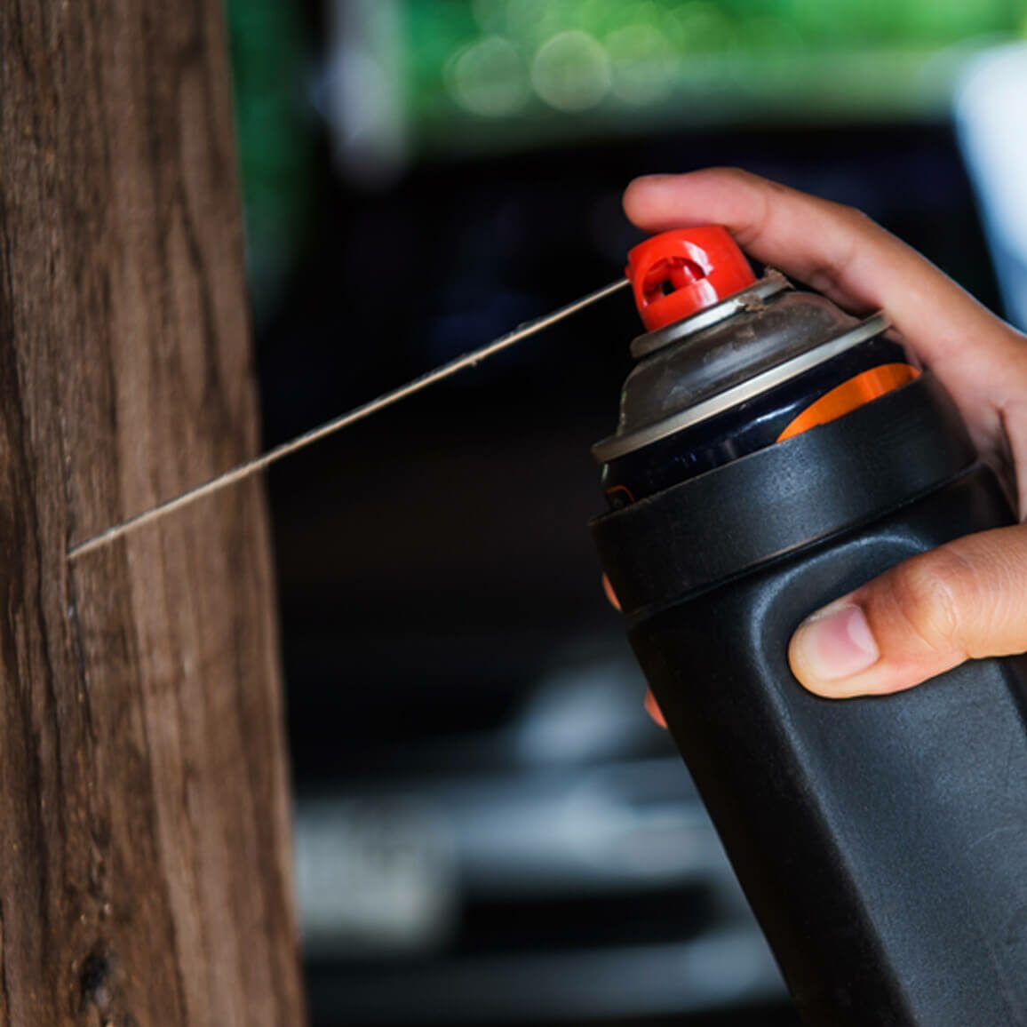 A person presses a spray can against a wooden surface, releasing a fine line of spray, with blurred vehicles in the background.