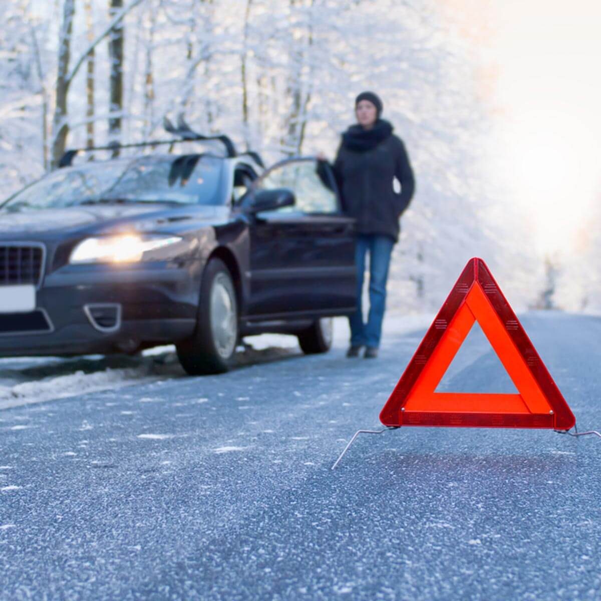 A triangular hazard sign is positioned on icy road asphalt, while a person stands beside a parked car, surrounded by snow-covered trees.