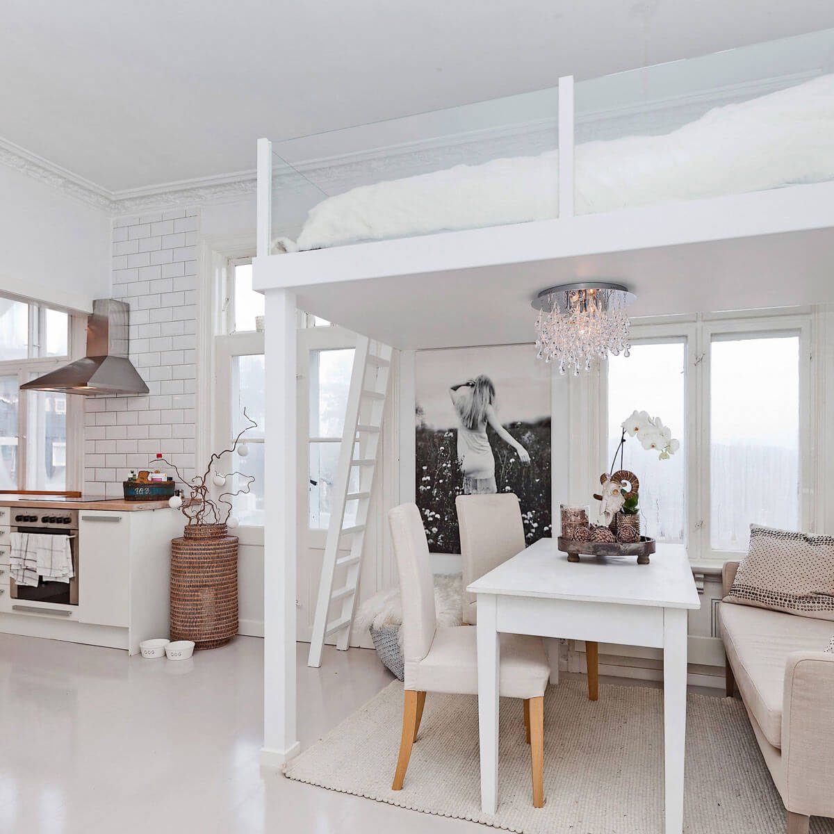 A white dining table sits with chairs in a bright room, featuring a lofted bed above, surrounded by windows and modern decor.
