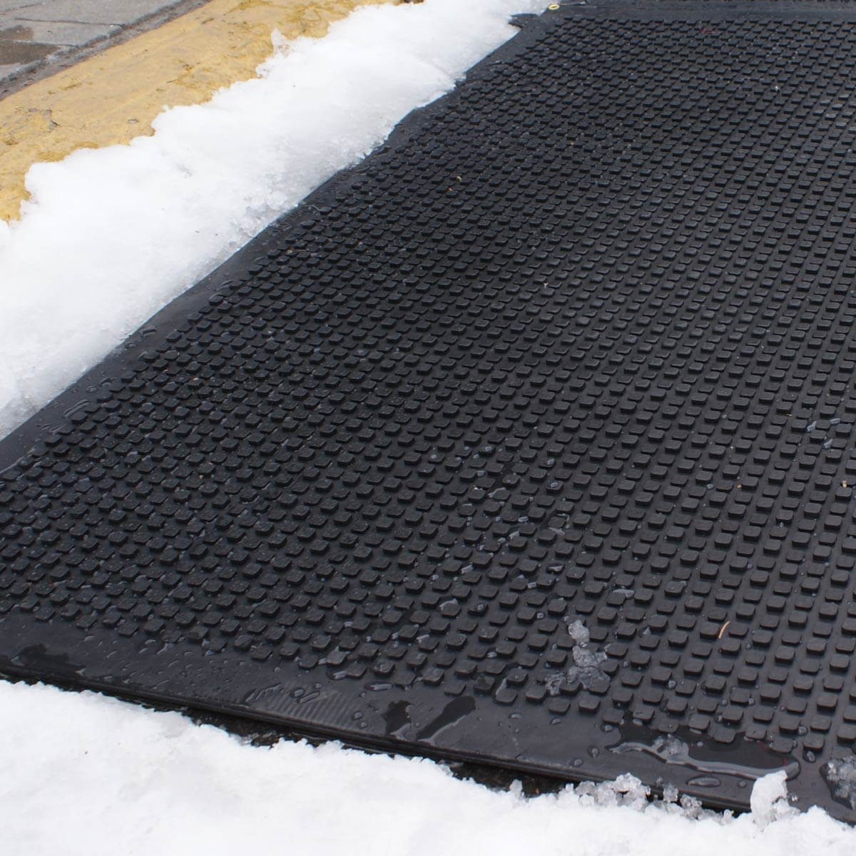 A black rubber mat sits on a snowy surface, retaining some water and ice, providing traction in a slippery environment.