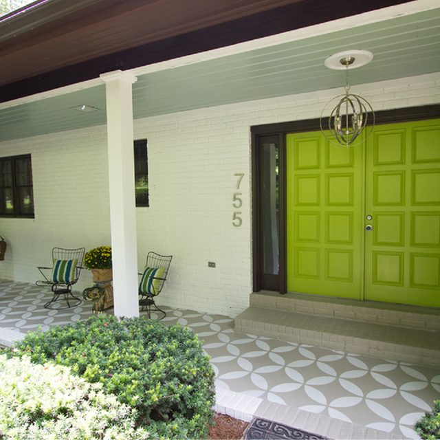 A vibrant green door stands open, inviting visitors onto a patterned porch surrounded by shrubs and two wire chairs, with house number 755 displayed nearby.
