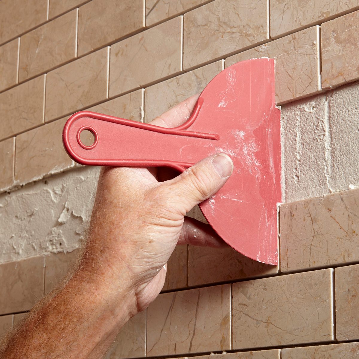 A hand holds a red plastic trowel, applying material to gaps between beige tile pieces on a wall during a tiling project.