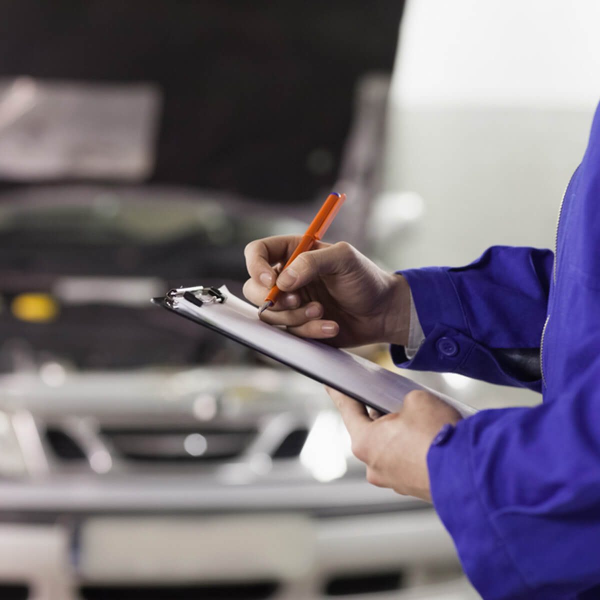 A mechanic holds a clipboard and an orange pen, recording information in a garage with a car's hood open in the background.