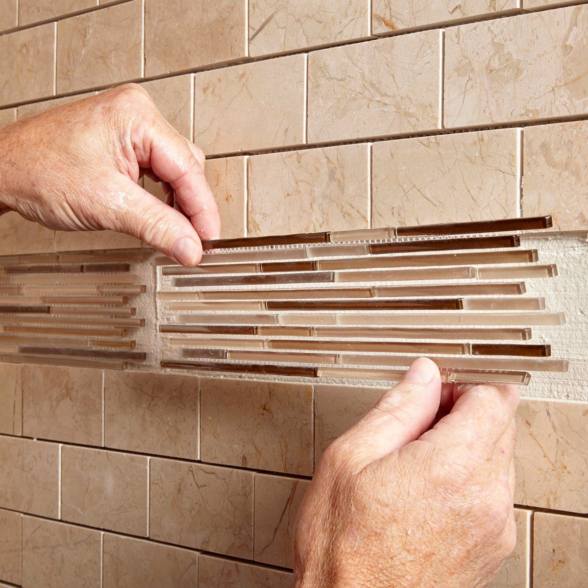 Hands are installing a decorative tile strip on a wall, surrounded by small beige tiles, in a likely bathroom setting.