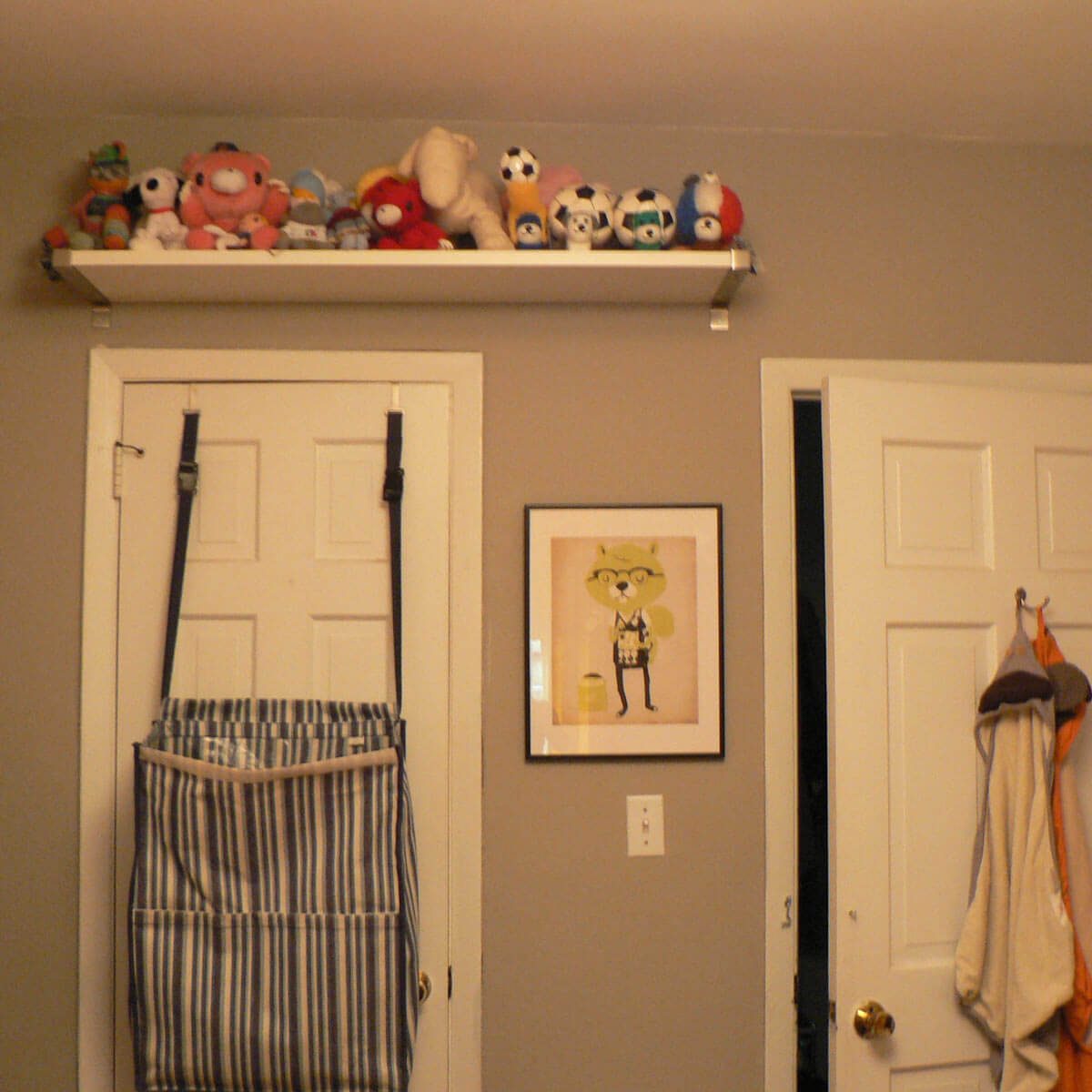 A shelf holds various stuffed animals above a door; a striped bag hangs on the door, adjacent to a framed cartoon character illustration.