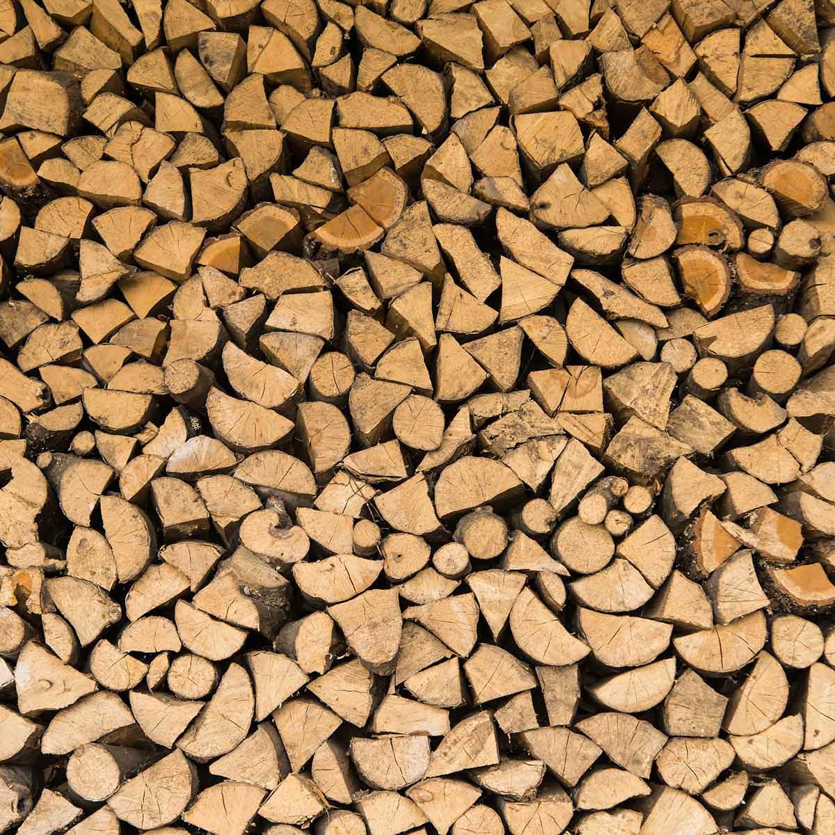 Stacked firewood, consisting of variously cut logs, fills the frame, creating a textured pattern of light brown surfaces and angular shapes in a rustic setting.