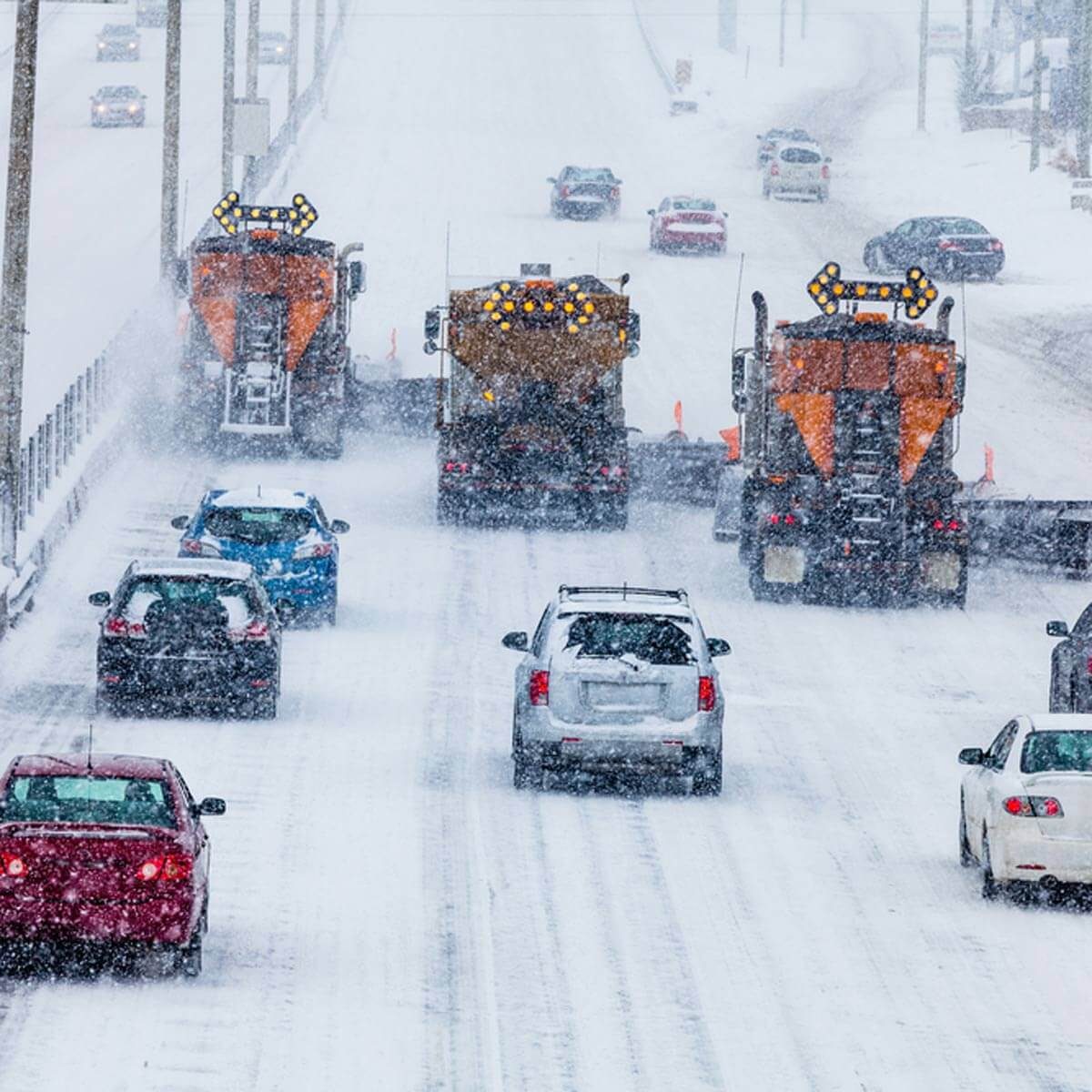 Two snow plows clear a snowy highway while cars navigate cautiously behind them in a heavy snowfall, creating a wintry, challenging driving environment.