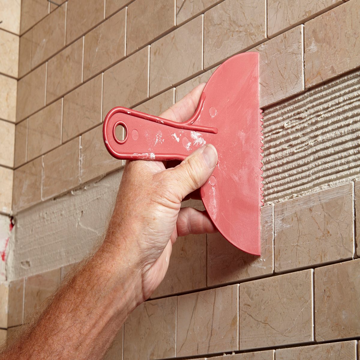 A hand holds a notched trowel applying mortar to a wall, surrounded by laid ceramic tiles, indicating a tiling process in a construction or renovation setting.