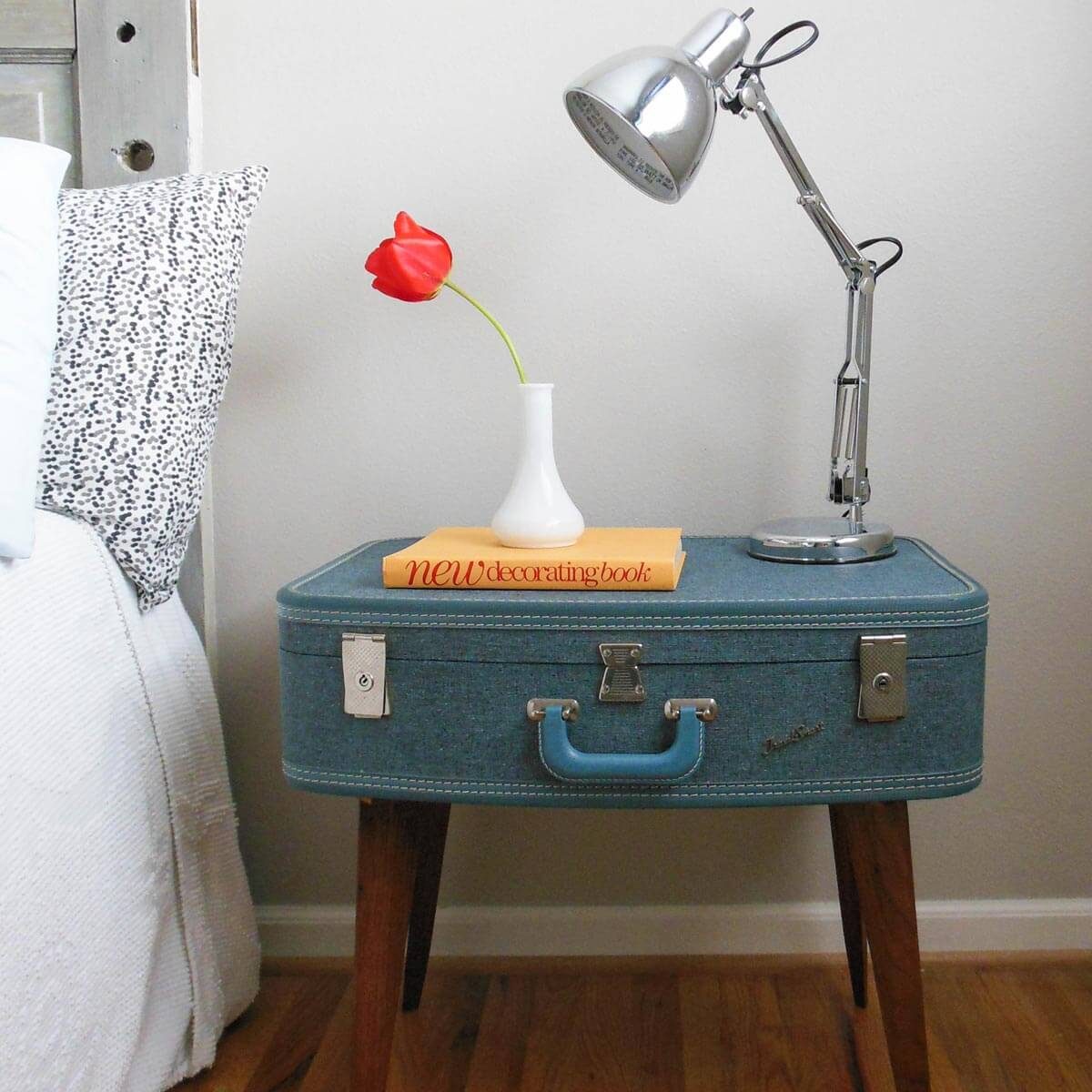 A vintage suitcase table holds a lamp, a small vase with a red tulip, and a book titled "new decorating book" in a cozy bedroom setting.