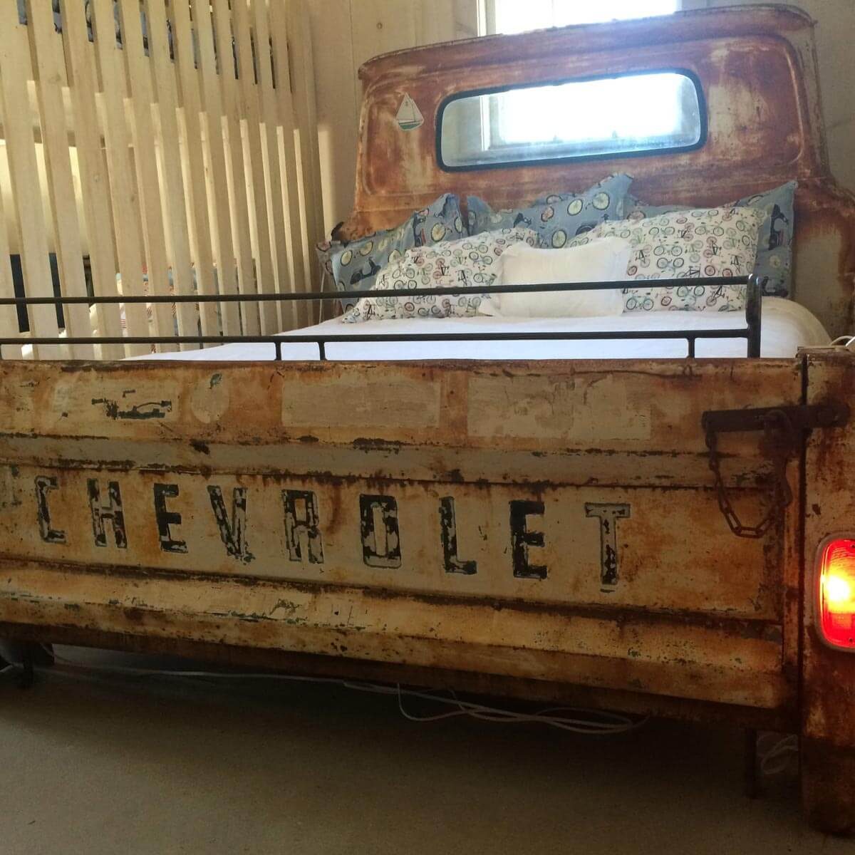 A vintage Chevrolet truck bed serves as a bed frame, adorned with colorful pillows and a white mattress, set against a rustic interior.
