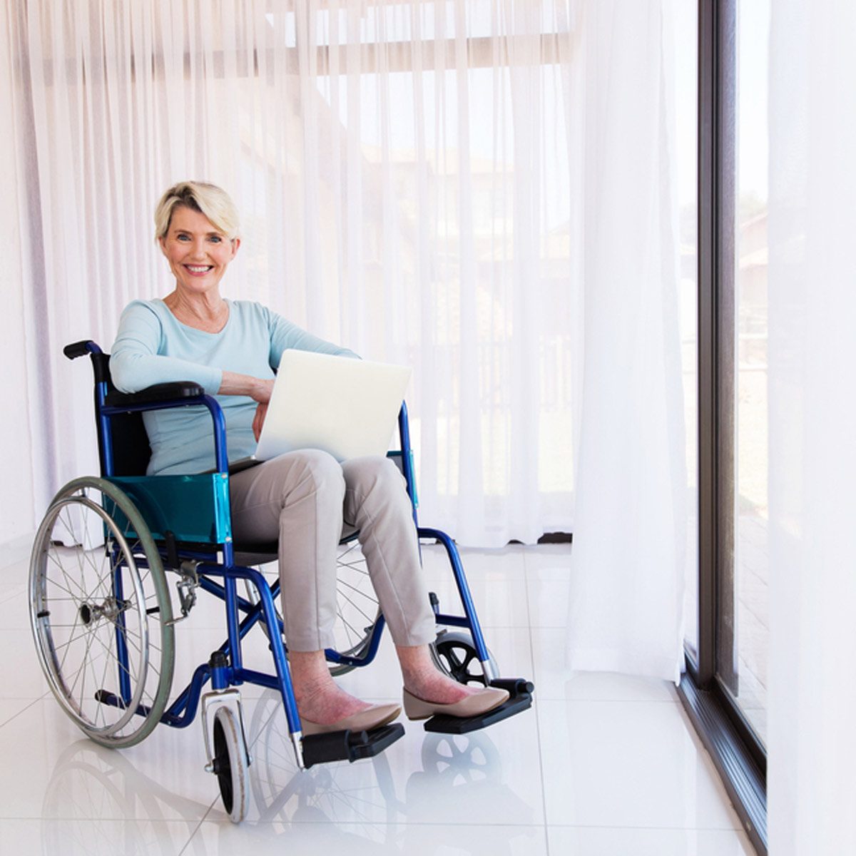 A woman in a wheelchair is using a laptop while sitting indoors, smiling. She is surrounded by sheer curtains and a bright, light-filled space.
