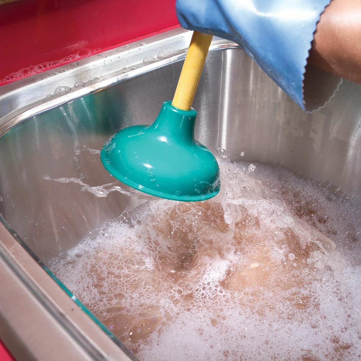 A green plunger is being used to agitate soapy water in a stainless steel sink, indicating an attempt to unclog it.