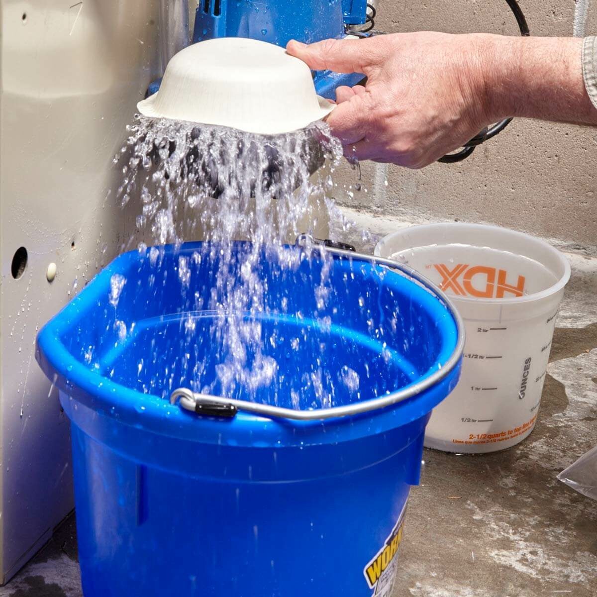 A hand tilts a white bowl, pouring water into a blue bucket below, surrounded by a concrete floor and a measuring cup nearby.