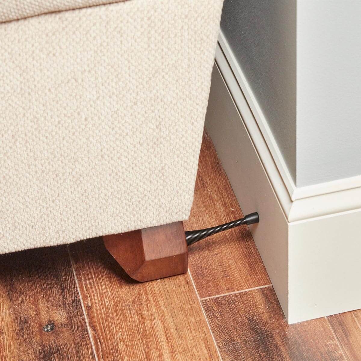 A couch leg is positioned on a wooden floor, with a metal screw adjusting its height against a wall