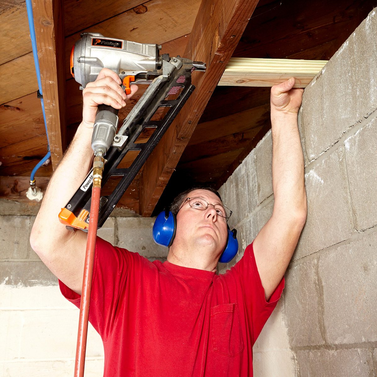 A man uses a nail gun to attach wood to a ceiling while wearing ear protection in a basement with exposed concrete walls.