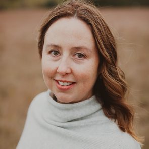 A woman with long, wavy brown hair smiles warmly, wearing a light gray sweater. She stands in a field with blurred greenery in the background.