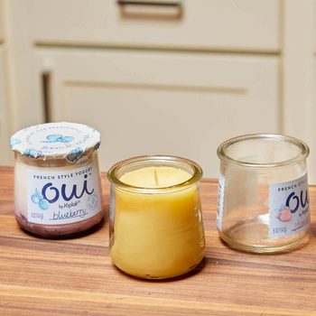 A blueberry yogurt container and a yellow candle sit on a wooden surface, with an empty yogurt jar nearby, against a light-colored background.