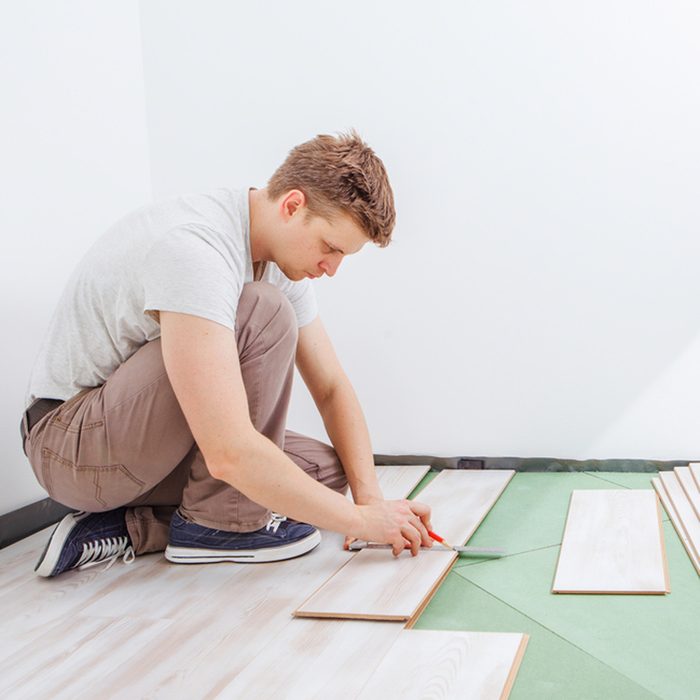 A man kneels on a green mat, measuring wooden floor planks with a ruler and cutting them, in a bright, minimalist room.