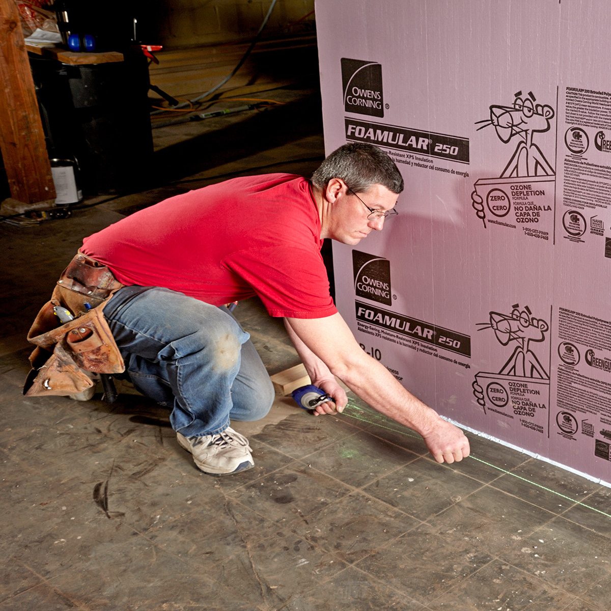 A man kneels on a floor, using a tool to mark a line on a foam board installed on a wall in a construction setting.